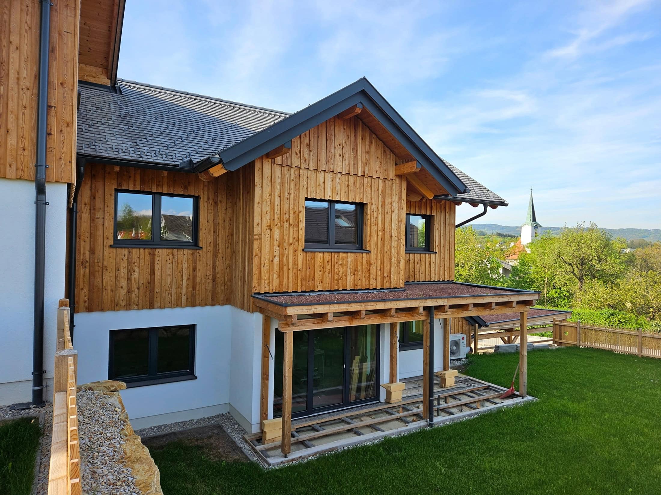 Modernes Holzhaus mit Pultdach, Garten und Terrasse im Bau, im Hintergrund eine Kirchturmspitze